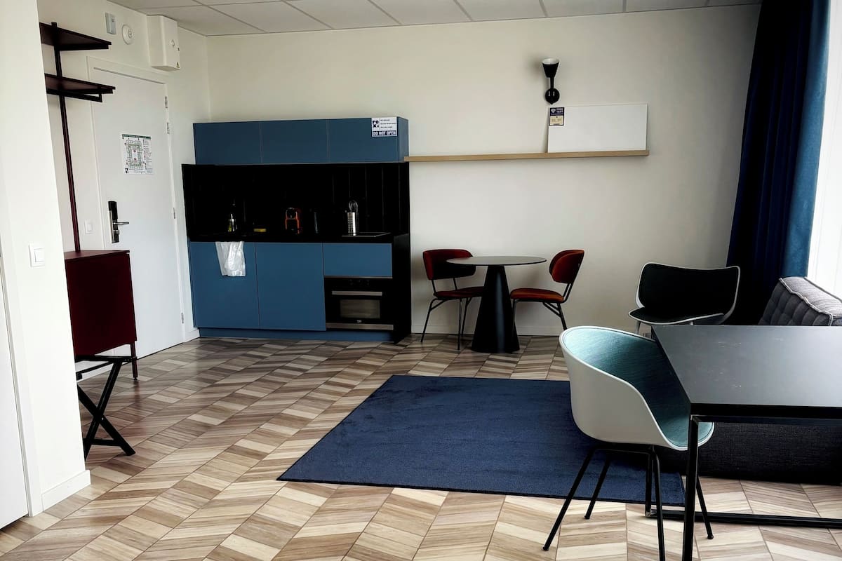A hotel room with a bed, a desk, a chair, a small table, a sofa, a blue rug, a TV, a shelf, and a wall-mounted air conditioner.