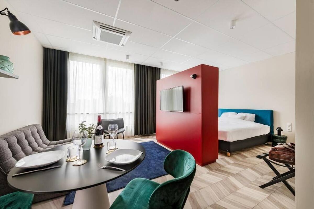 A hotel room with a sofa, dining table, and a bed with a red accent wall.