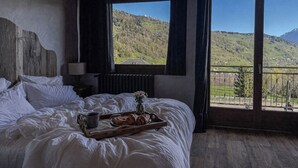 Un dormitori amb un capçal de fusta, un llit amb roba de llit blanca, una bandeja amb pastissos i una tassa, i una vista d'un paisatge verd a través de grans finestres.