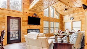 Un interior de cabaña con detalles de madera, un televisor en la pared, un sofá, un sillón reclinable y una mesa de comedor con sillas.