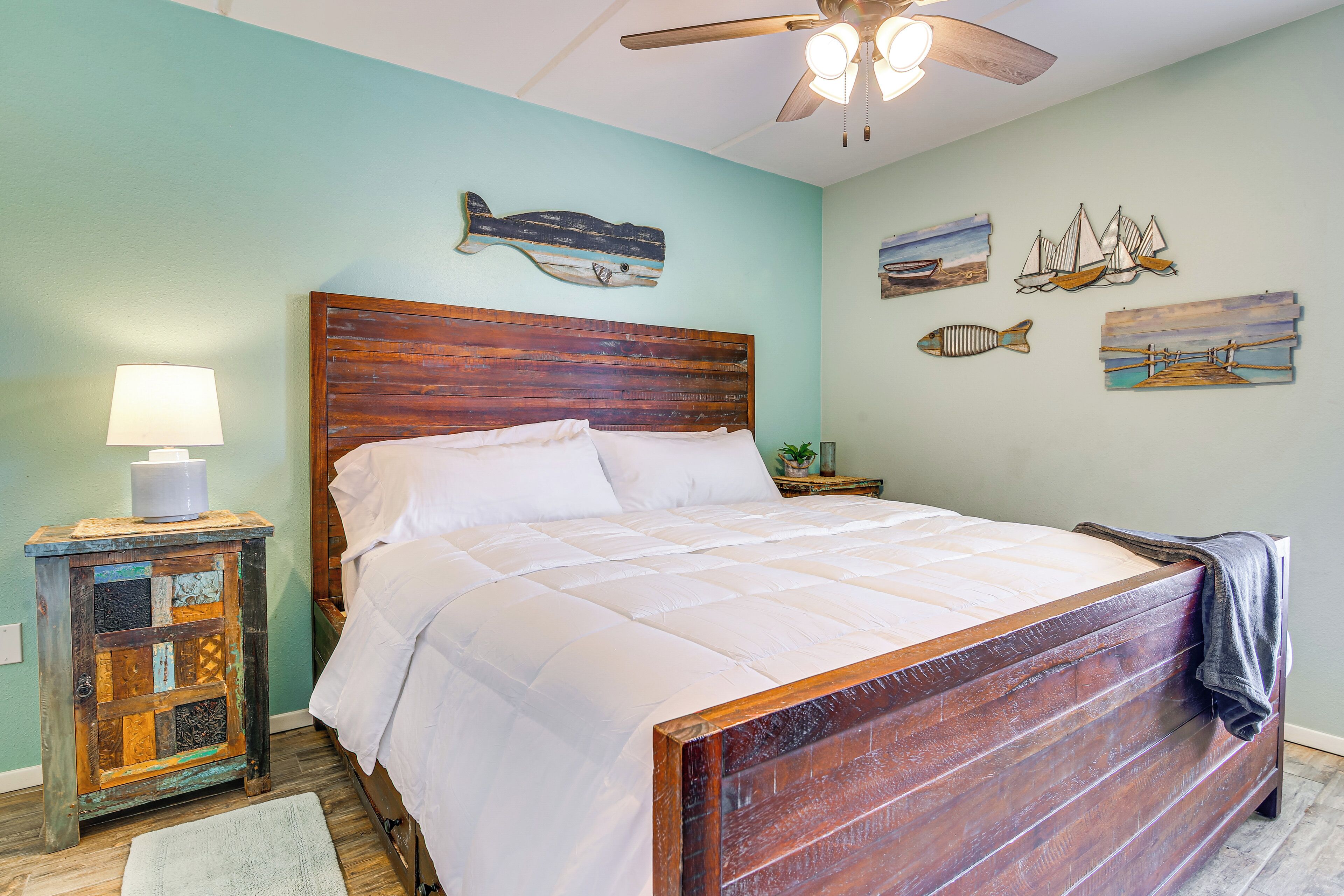 A bedroom with a wooden headboard, a bed with white bedding, a nightstand with a lamp, and wall art depicting fish and boats.