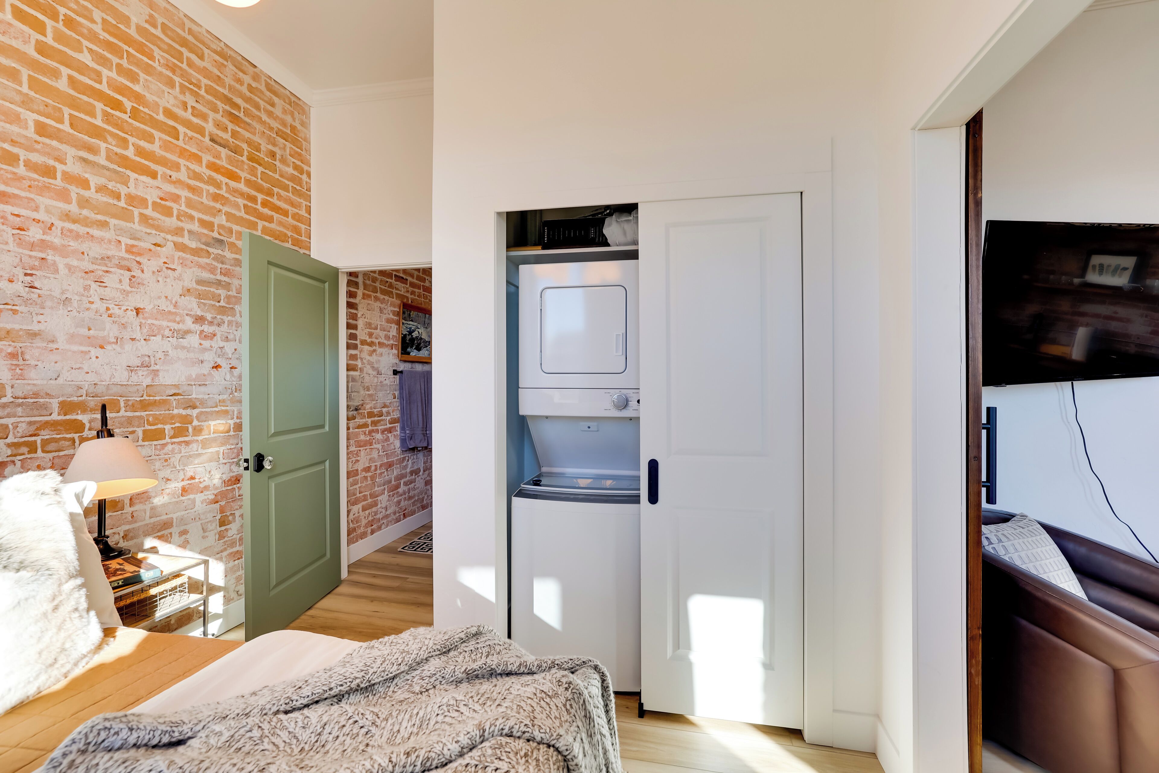 A compact room with a brick wall, a bed with a fluffy blanket, a small table with a lamp, and a white washing machine in the corner.