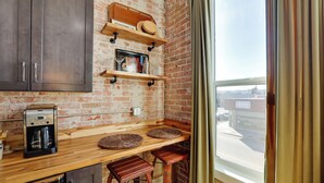 A kitchen with a brick wall, wooden shelves, a coffee maker, and two stools. - Walk to Shops & Eats: Charming Dtwn Emmett Apt! (Emmett)