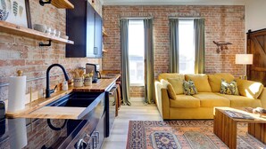 A cozy living room with a yellow sofa, a wooden coffee table, and a kitchen area with a black sink and wooden shelves. - Walk to Shops & Eats: Charming Dtwn Emmett Apt! (Emmett)