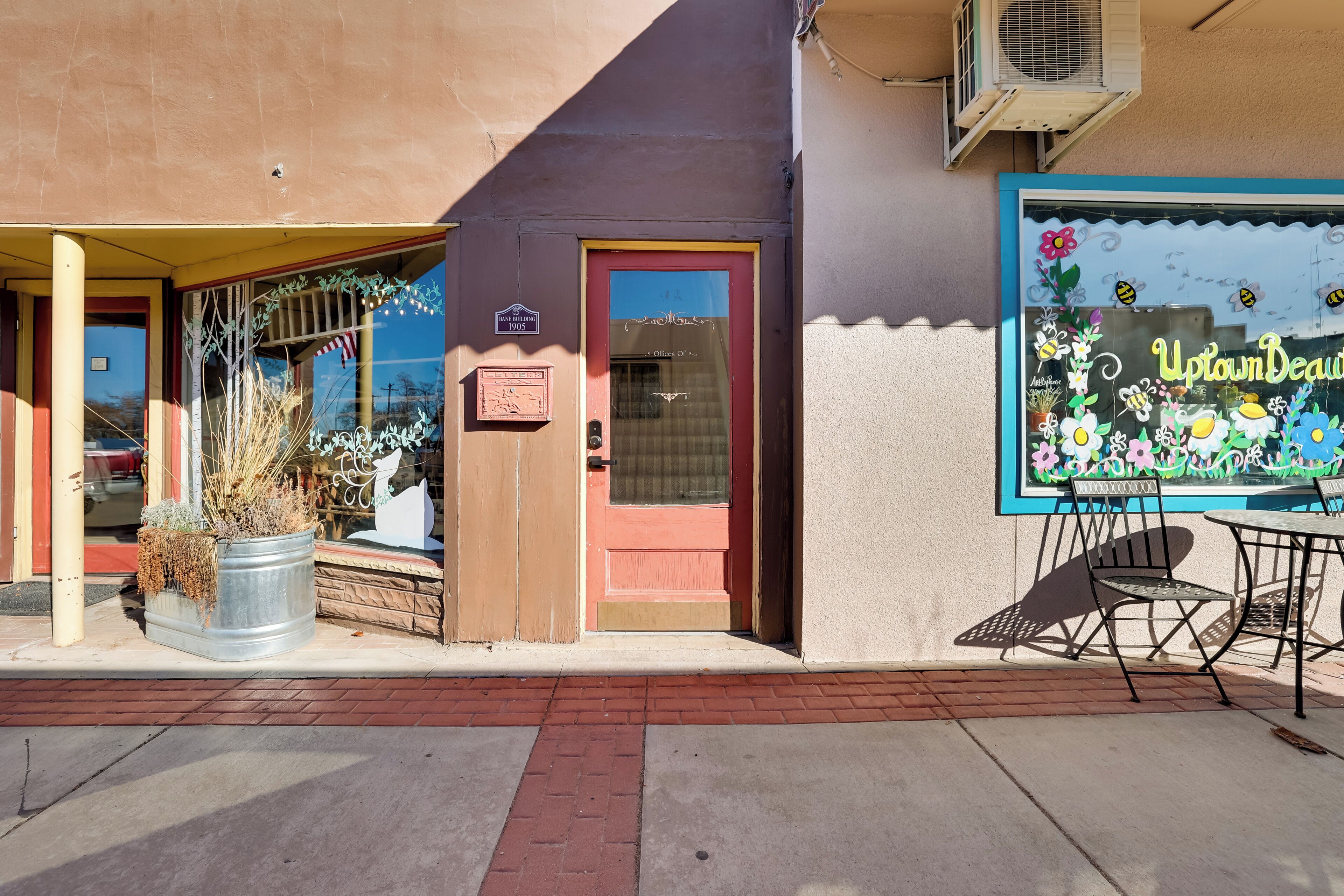 A storefront with a red door, a window with a floral design, and a metal chair outside.