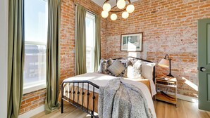 A bedroom with a brick wall, a bed with a headboard, a bedside table with a lamp, and a window with curtains. - Walk to Shops & Eats: Charming Dtwn Emmett Apt! (Emmett)
