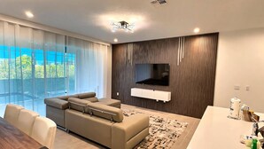 A modern living room with a sectional sofa, a flat-screen TV mounted on a wooden panel, a dining area with a white table, and a large window with a view of greenery. - Luxury 6BR Pool Home at Solara Resort (Kissimmee)