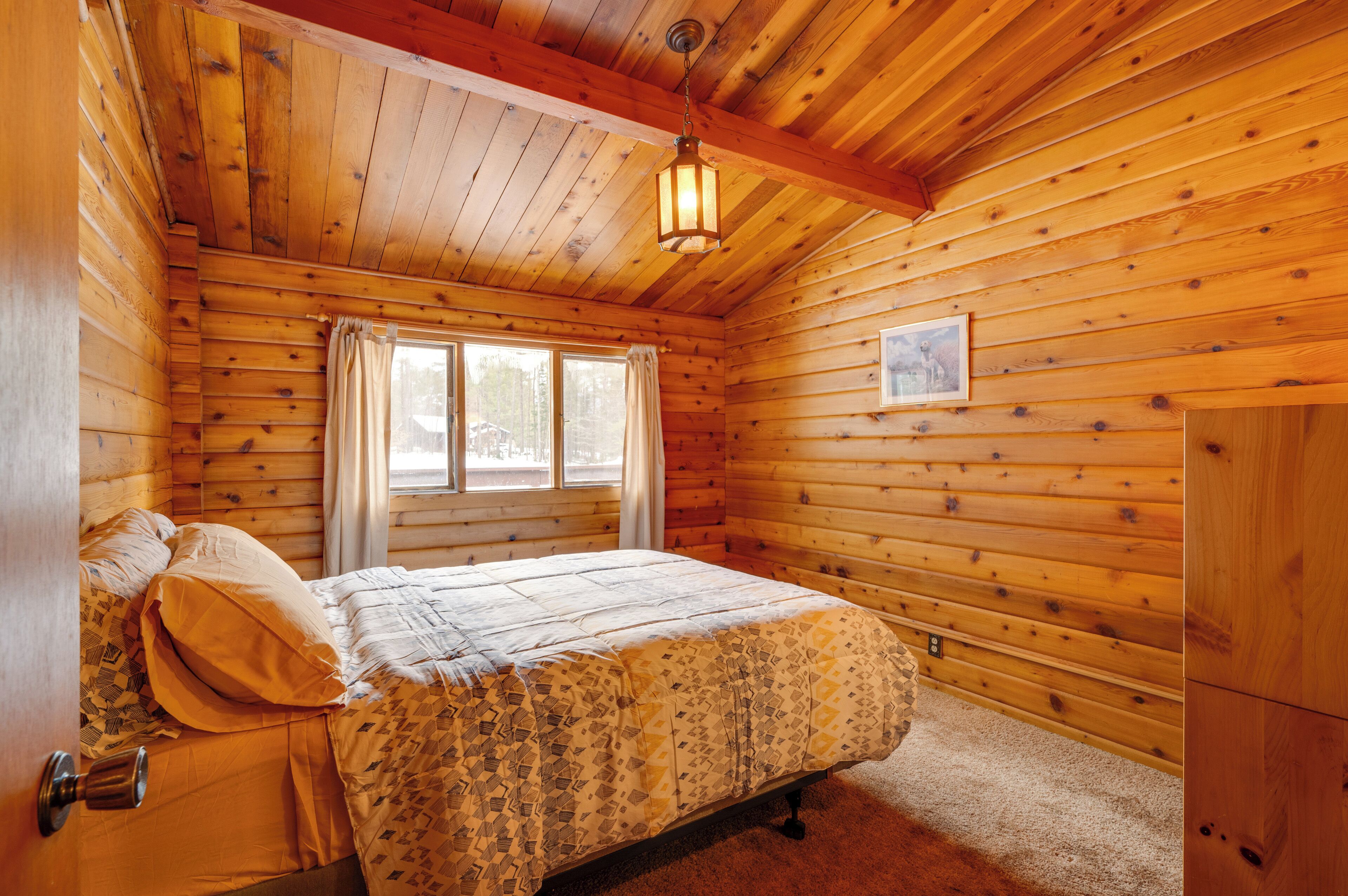 A wooden cabin bedroom with a bed, a window with curtains, a wooden ceiling, and a framed picture on the wall.