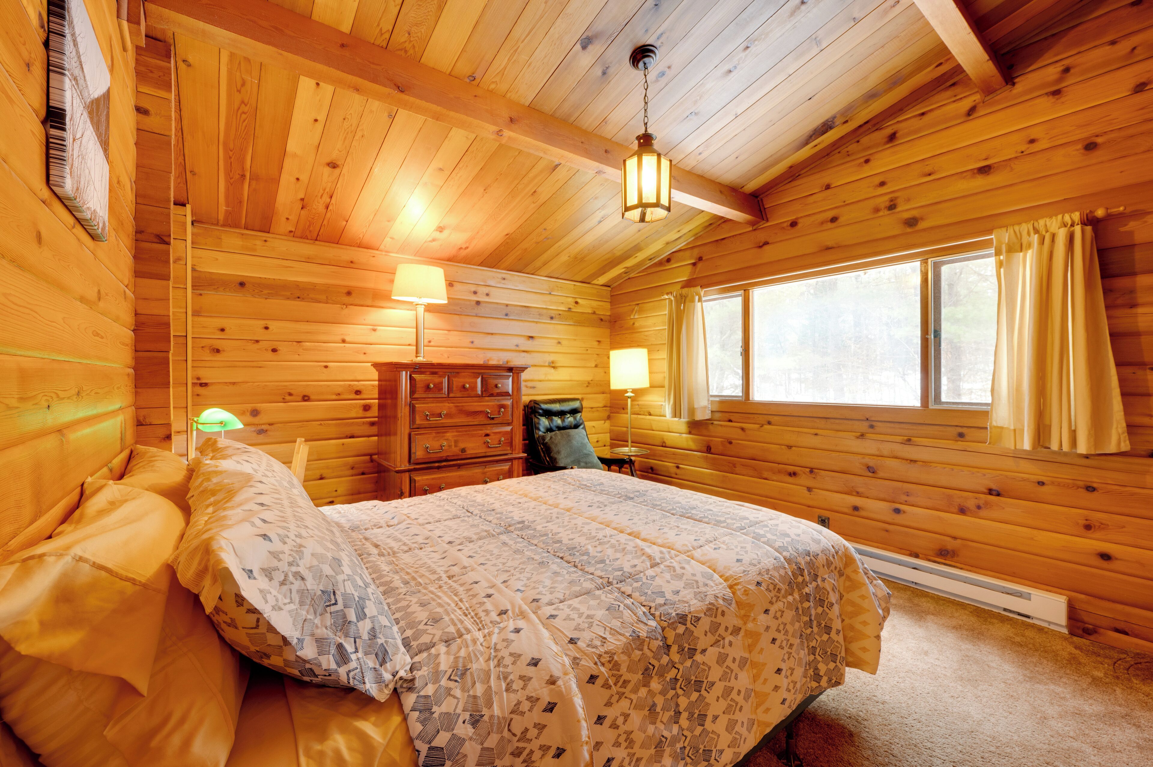 A wooden cabin bedroom with a bed, two bedside lamps, a dresser, and a window with curtains.
