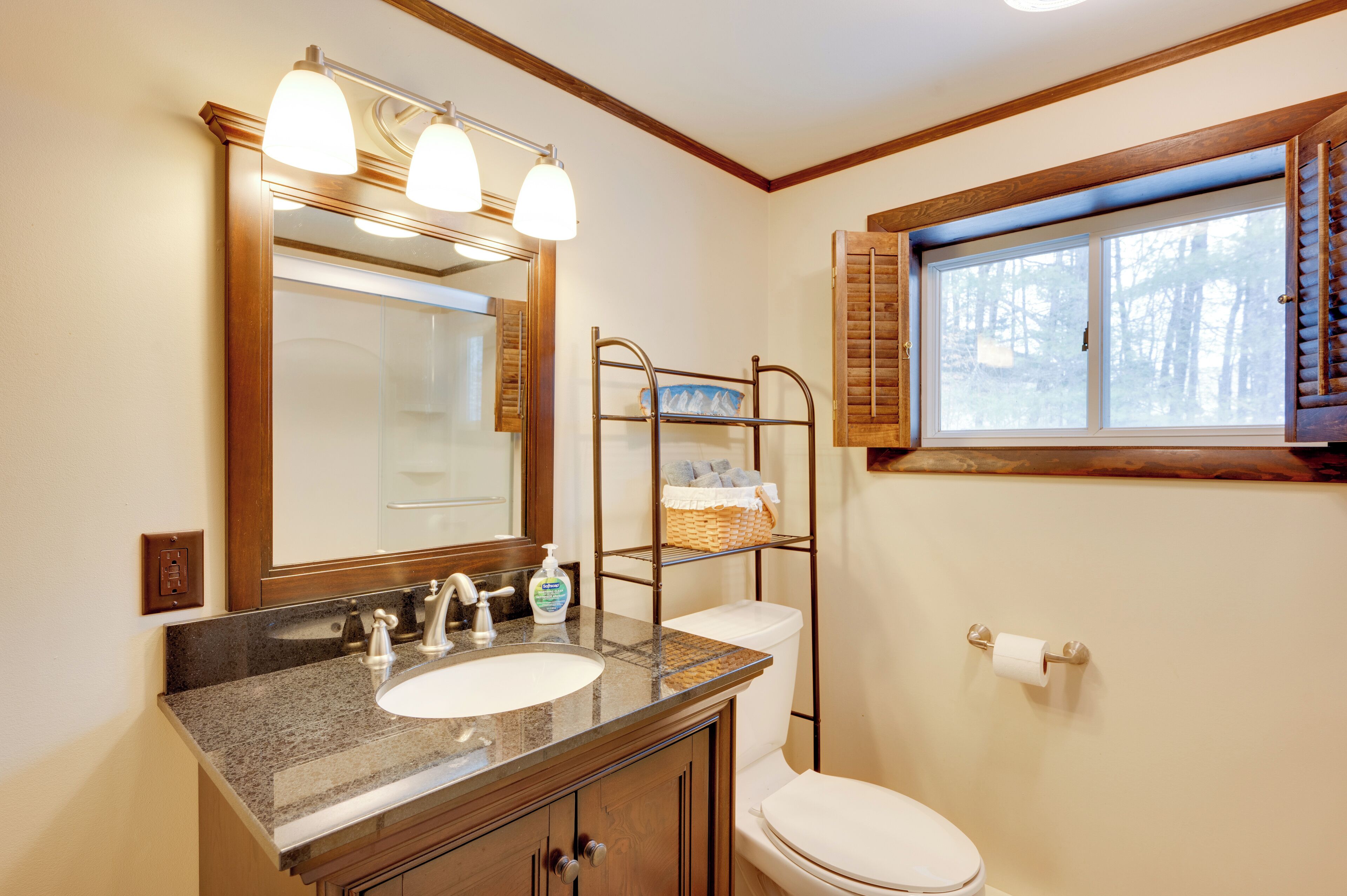 Une salle de bain avec un lavabo sur un plan de travail en granit, un miroir avec trois lampes, des toilettes et une fenêtre avec des volets.