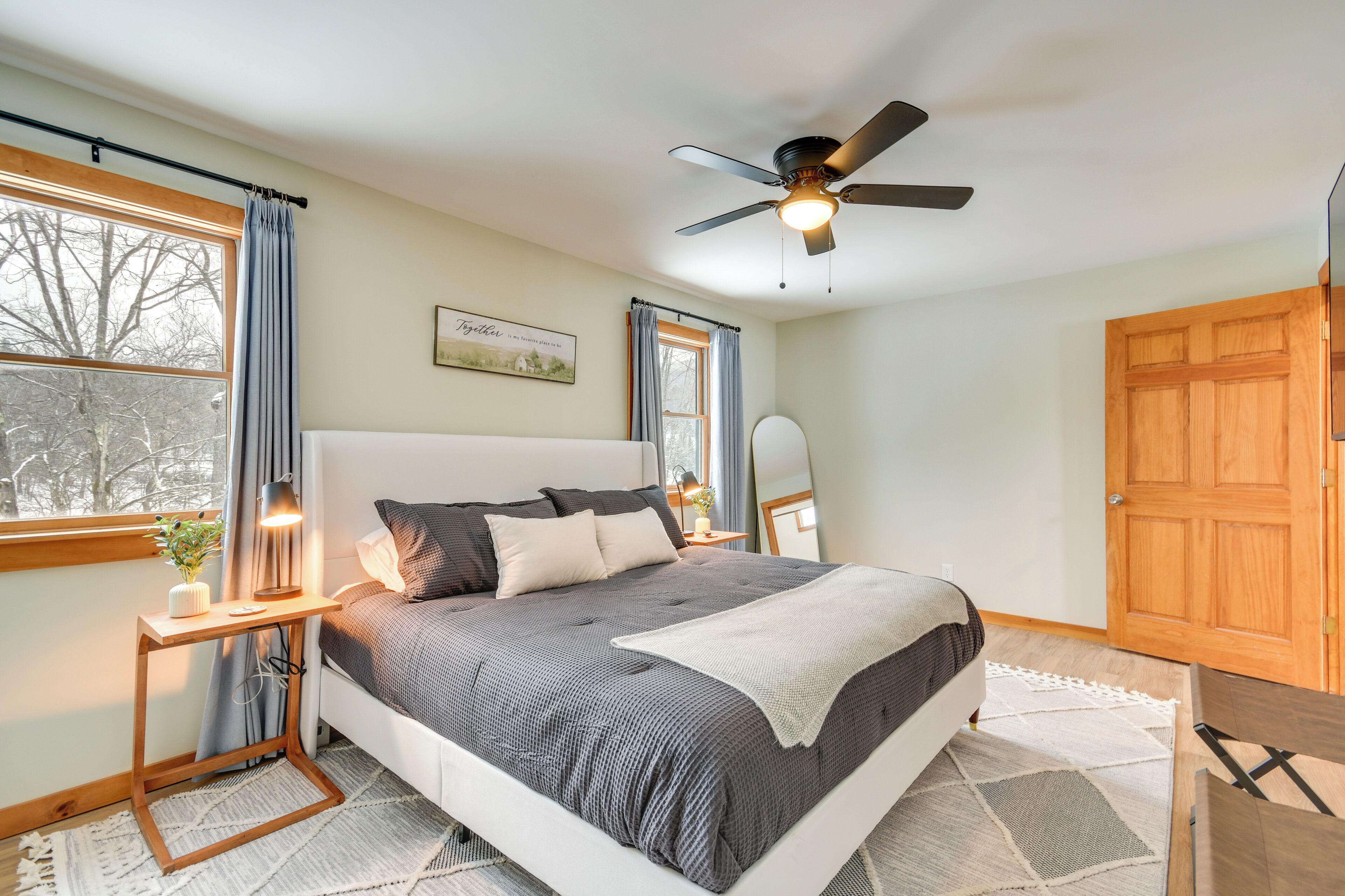 Un dormitorio con cama, mesitas de noche, ventilador de techo, ventana con cortinas y puerta de madera.