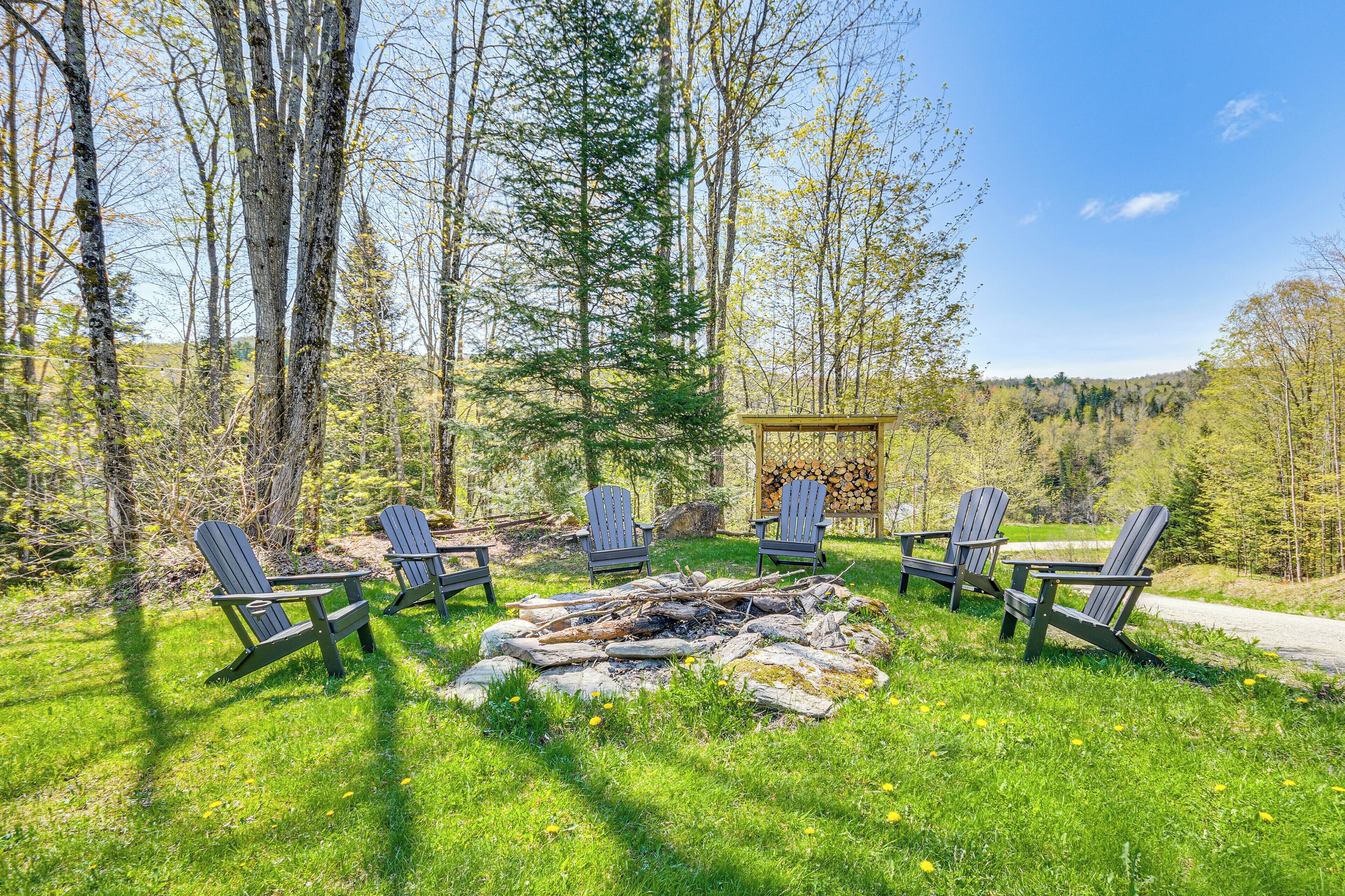 A fire pit with logs surrounded by Adirondack chairs in a forested area.