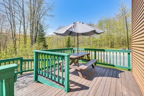 A wooden deck with a picnic table and umbrella, surrounded by a green railing, overlooking a forested area.