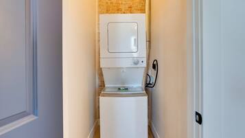 Una lavadora blanca y compacta en un pasillo estrecho con una pared de ladrillo y una puerta a la izquierda.