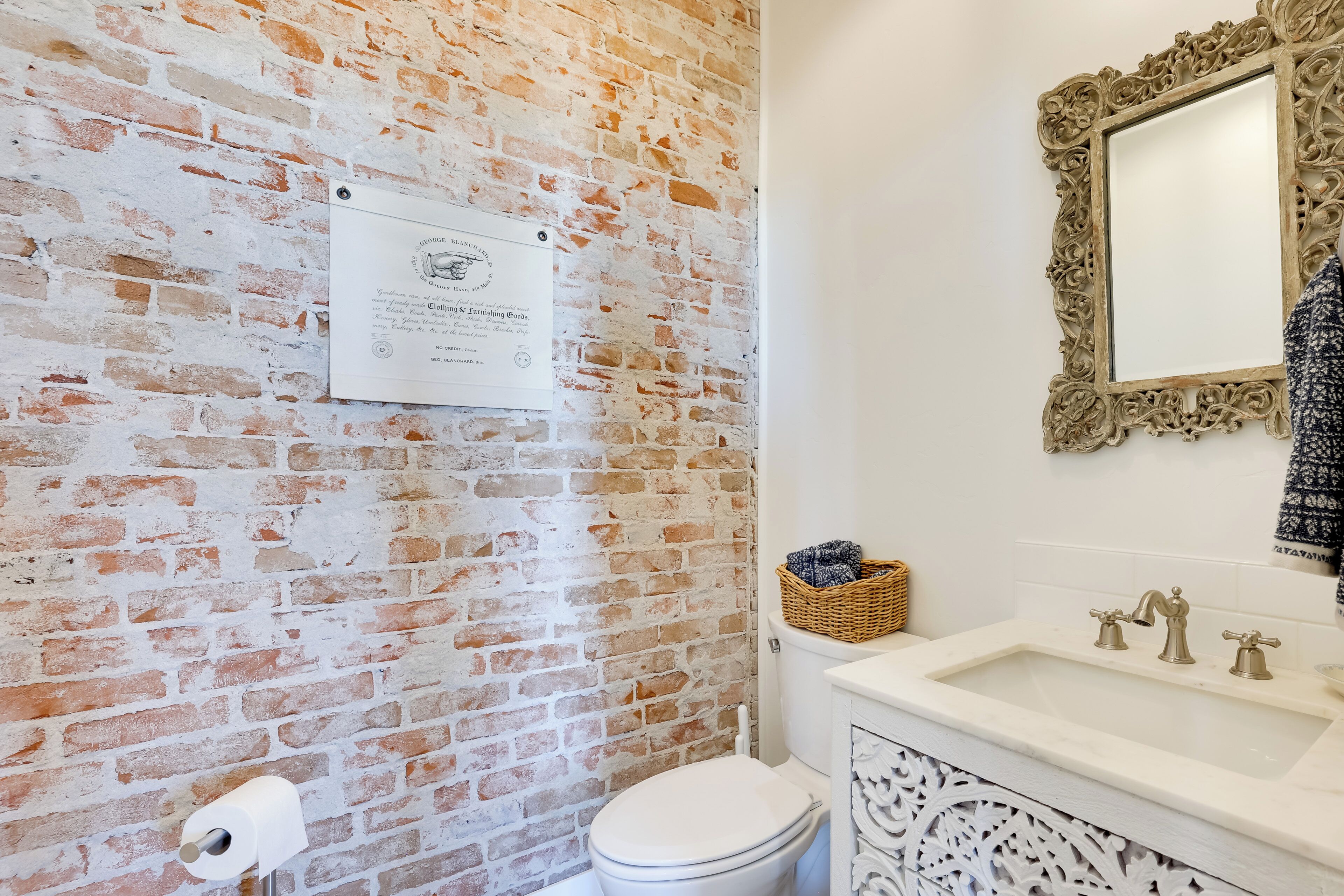 A bathroom with a brick wall, a toilet, a sink with a decorative mirror, and a wall-mounted plaque.