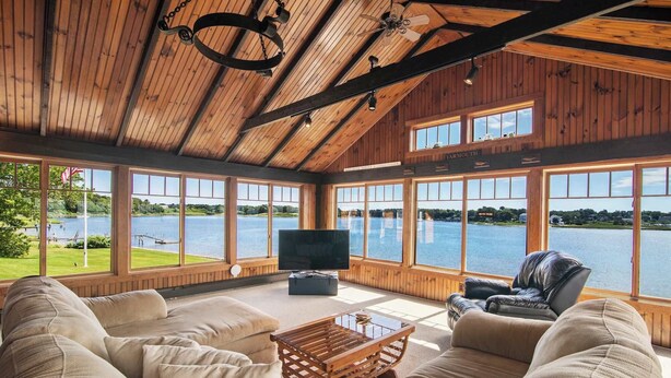 A spacious room with a large window overlooking a lake, a comfortable sofa, a leather chair, and a television. - Stunning Waterfront Home! (Yarmouth)
