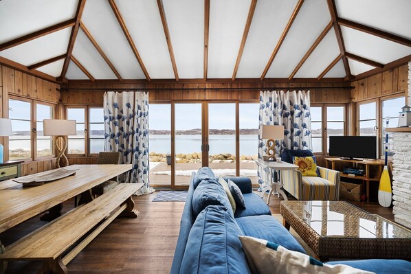 A cozy room with a wooden ceiling, large windows, a sofa, a table, and a TV. - Chick Coop (Suttons Bay)