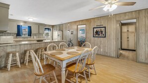 Un grand espace à manger avec une table en bois et des chaises blanches, une cuisine dotée d’un plan de travail blanc et d’un ventilateur de plafond.