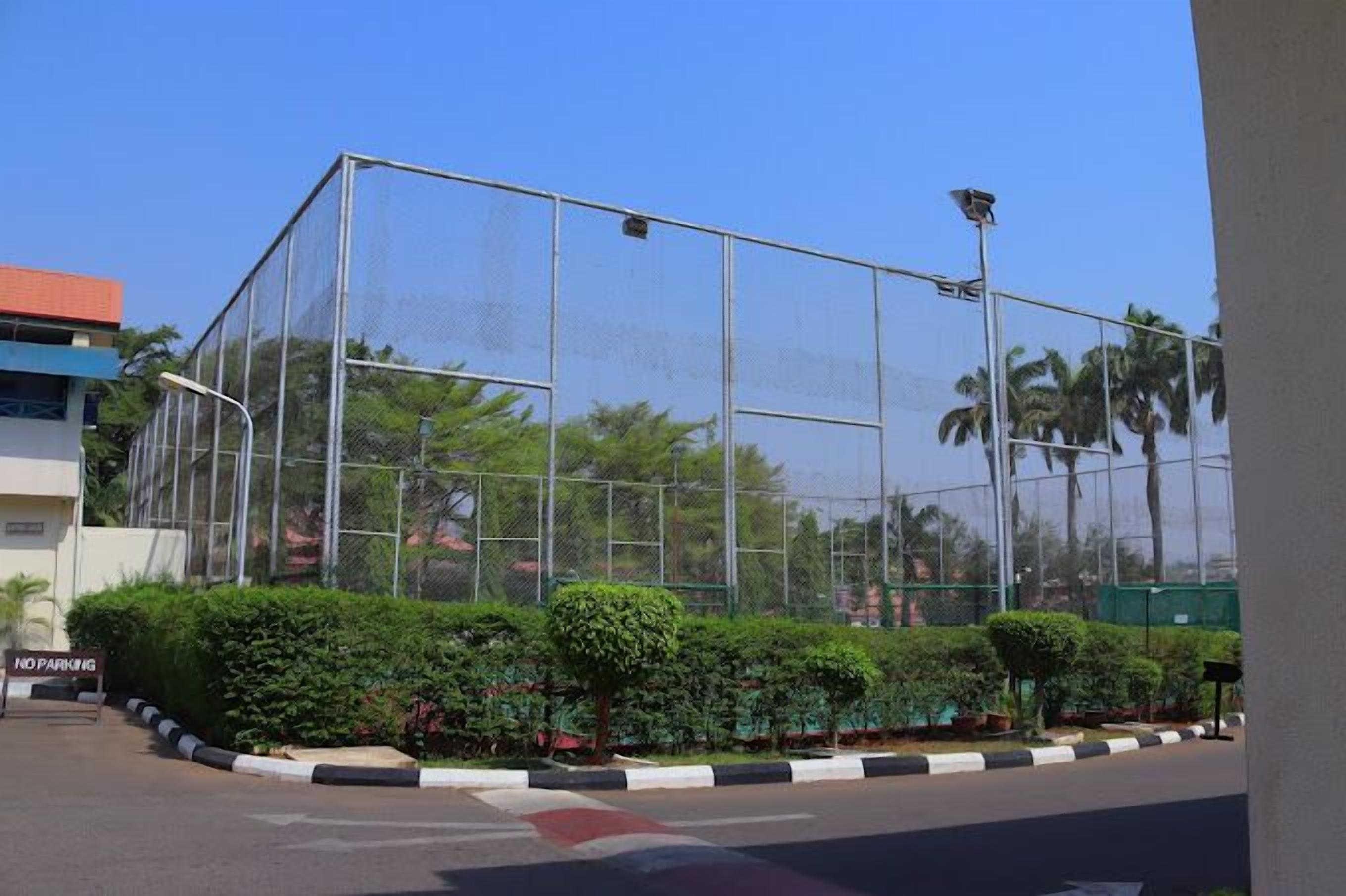 A fenced sports court with a "NO PARKING" sign, surrounded by well-maintained hedges and palm trees.