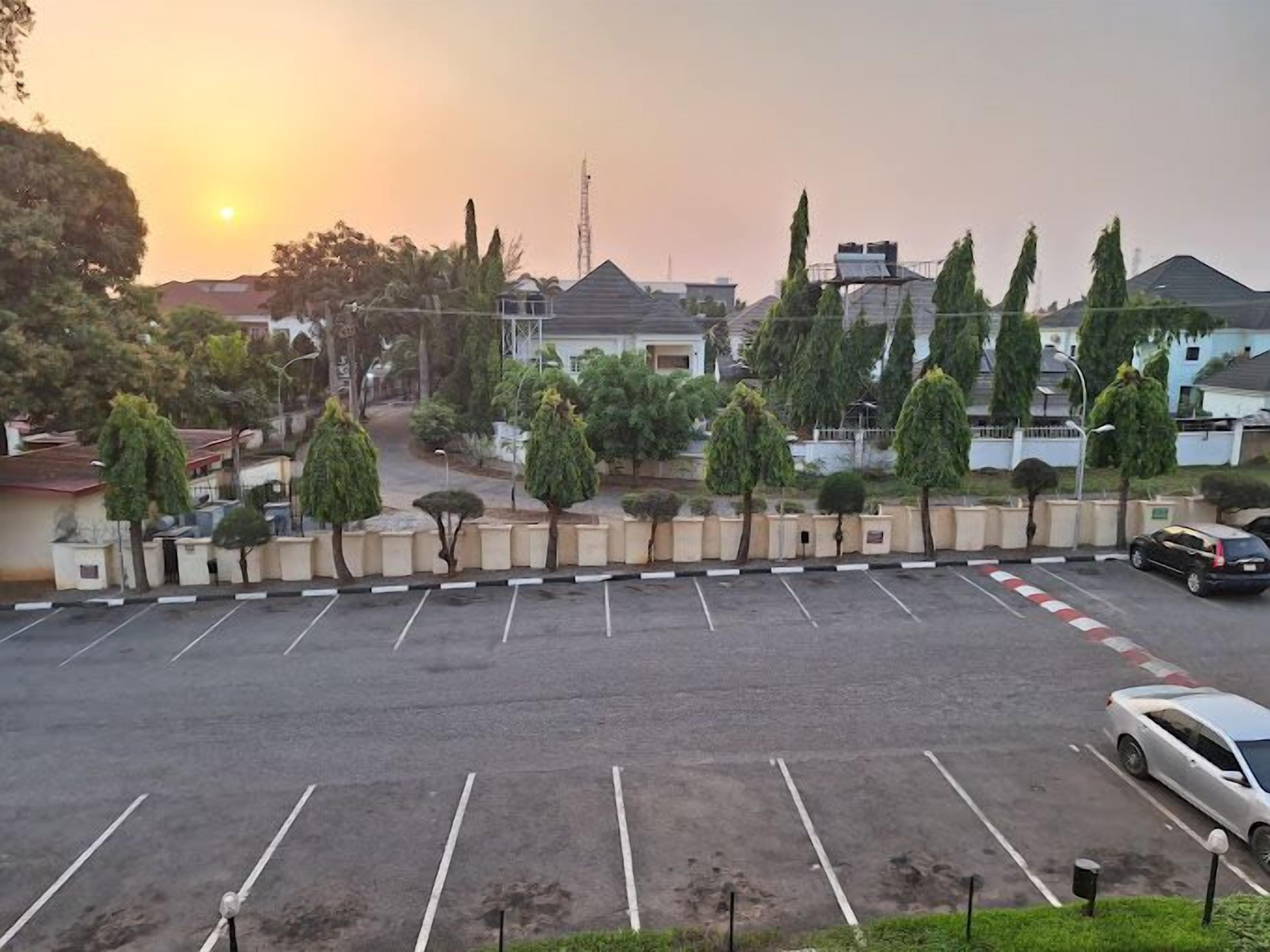 A parking lot with multiple parking spaces, a white car parked in one of the spaces, and a row of trees and buildings in the background.