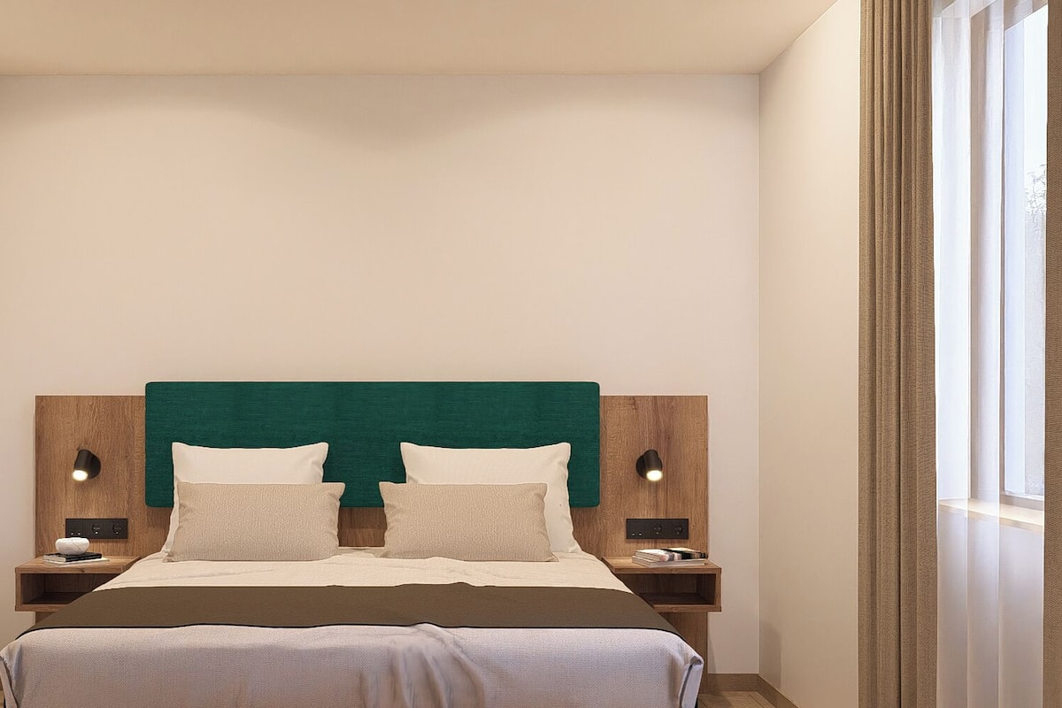 Une chambre à coucher moderne avec une tête de lit recouverte d’un tissu vert, deux tables de chevet avec des lampes et une grande fenêtre avec des rideaux.