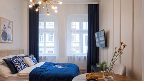 A hotel room with a bed, a round wooden table with a cup and pastries, a TV on the wall, and a window with curtains. - Neon 45 Apartment - ProperUNIT (Wroclaw)