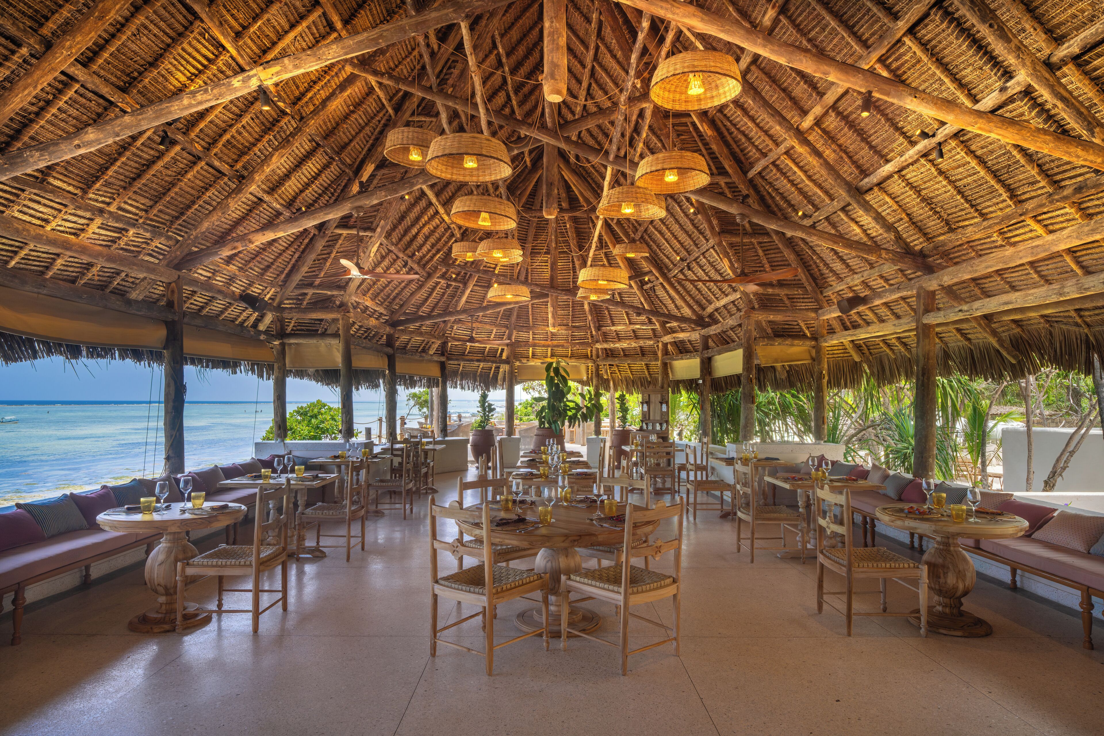Un amplio espacio al aire libre con mesas y sillas de madera, rodeado por una estructura de techo de paja.
