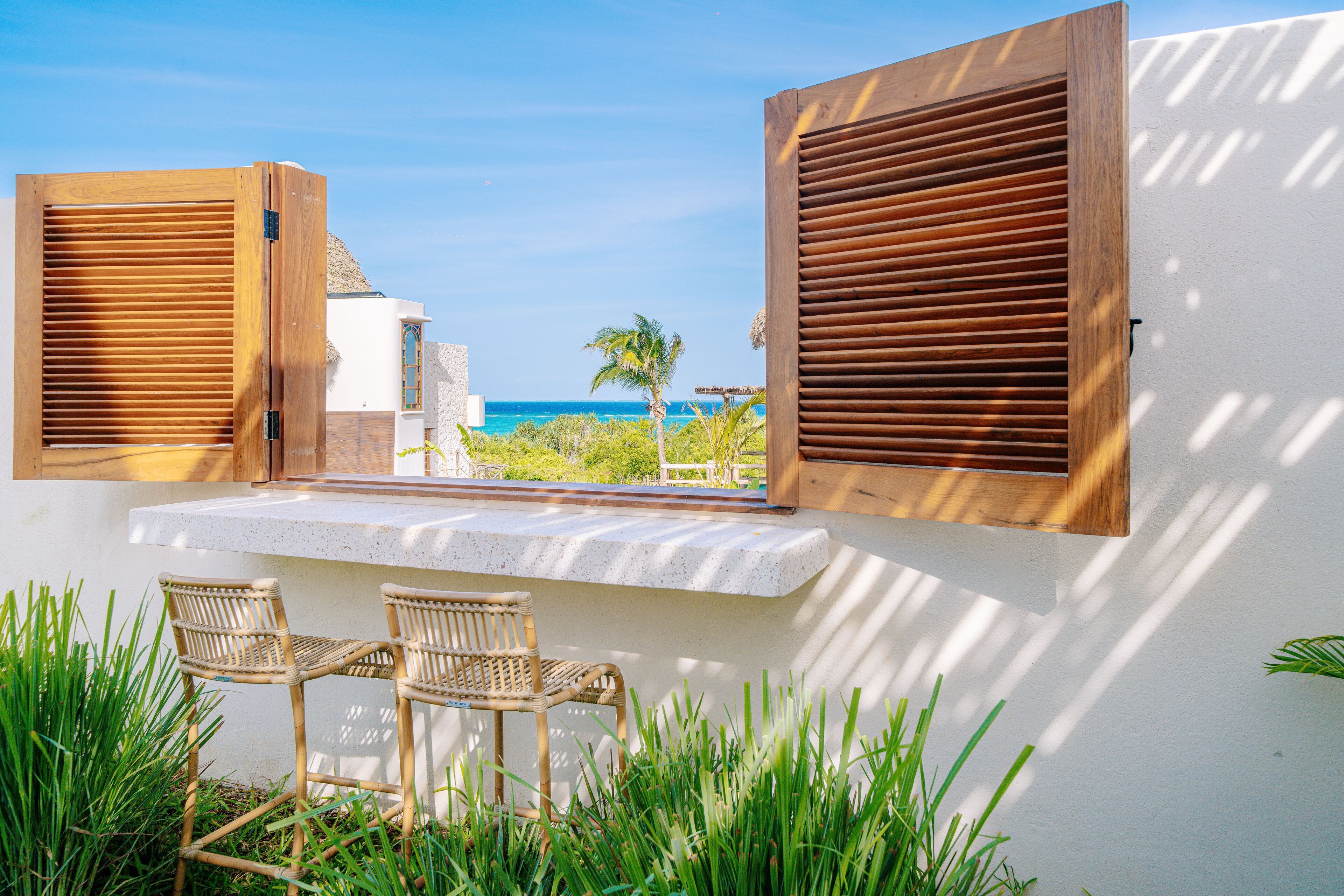 A balcony with wooden shutters, two wicker chairs, and a view of the ocean.