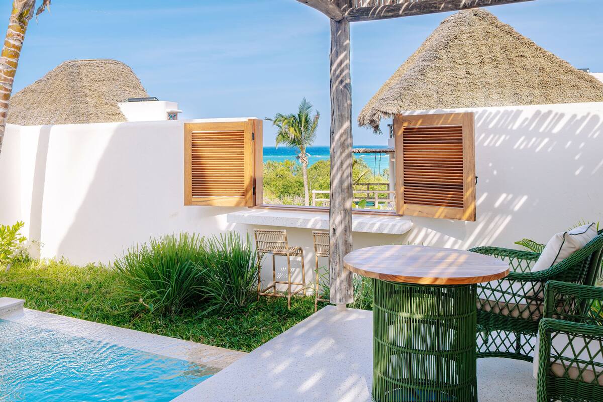 A poolside area with a view of the ocean, a thatched-roof structure, wicker furniture, and a small table.