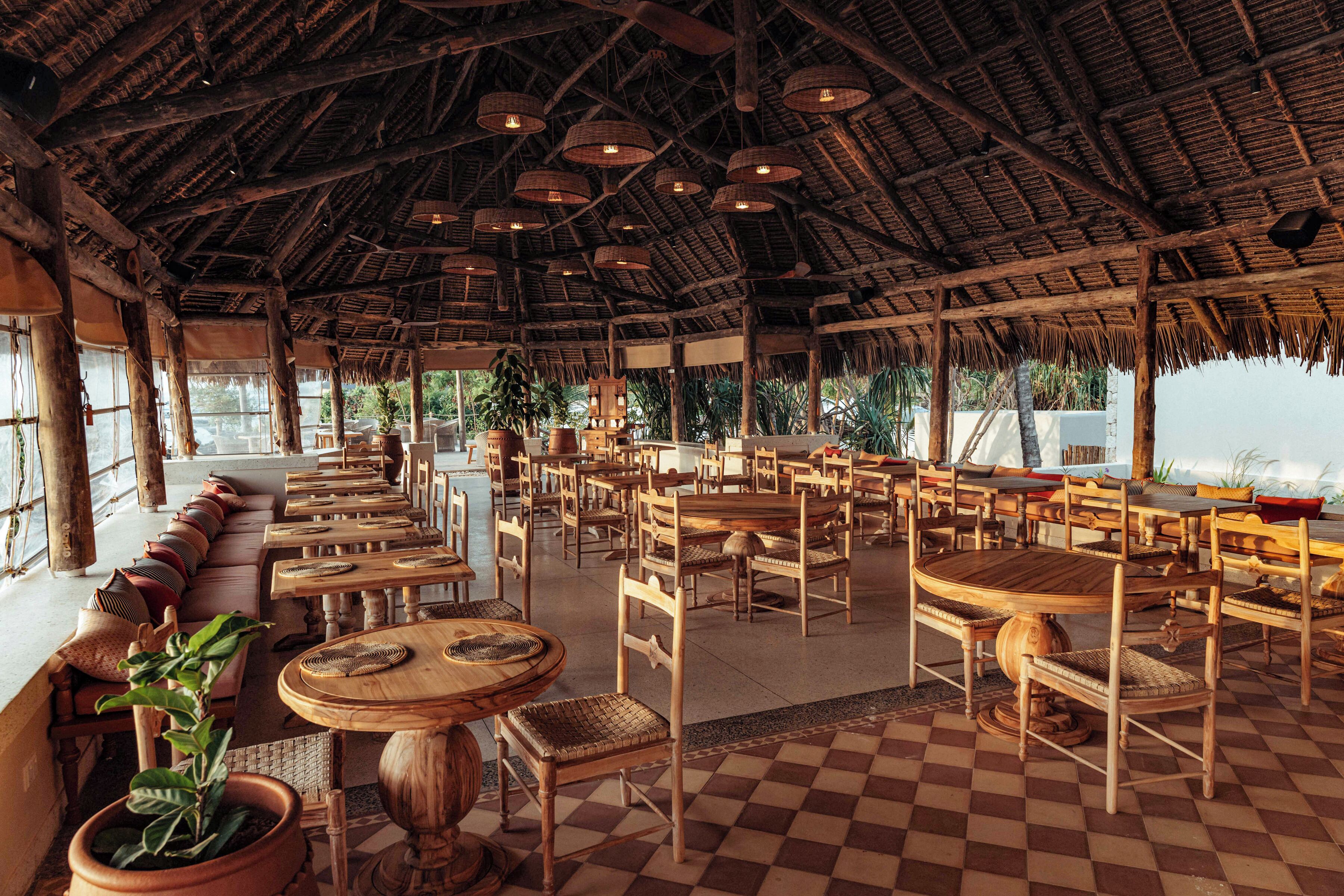 A spacious outdoor dining area with wooden tables and chairs, a thatched roof, and large windows overlooking a scenic view.