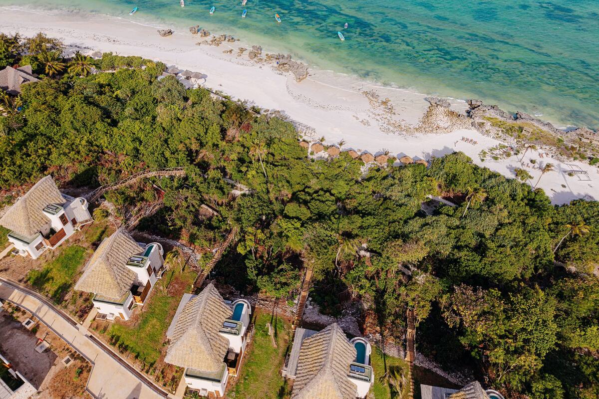 An aerial view of a coastal area with a beach, clear turquoise waters, and a cluster of houses with thatched roofs and a swimming pool.