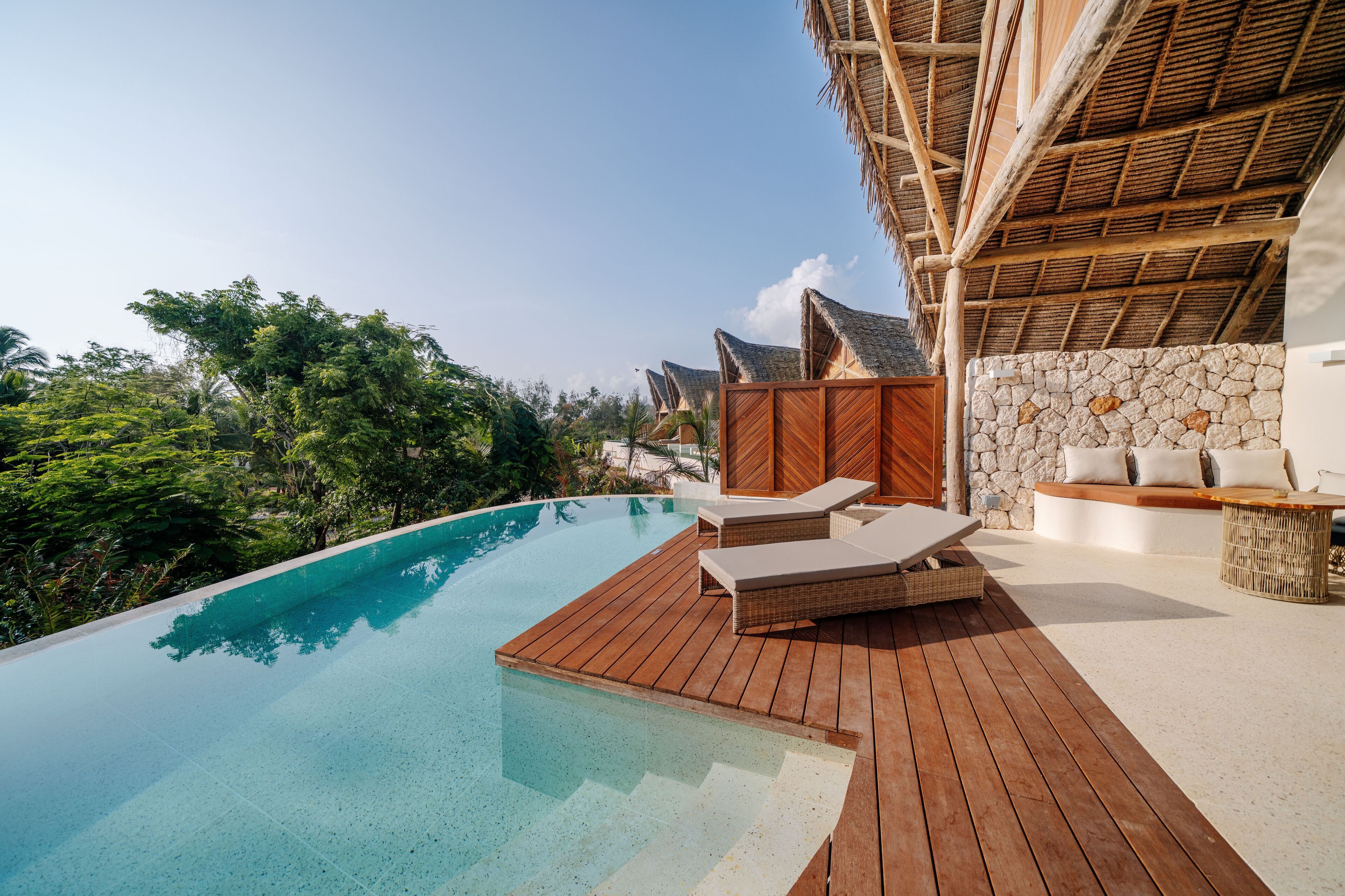 An infinity pool with wooden deck and lounge chairs, surrounded by lush greenery and a stone wall.