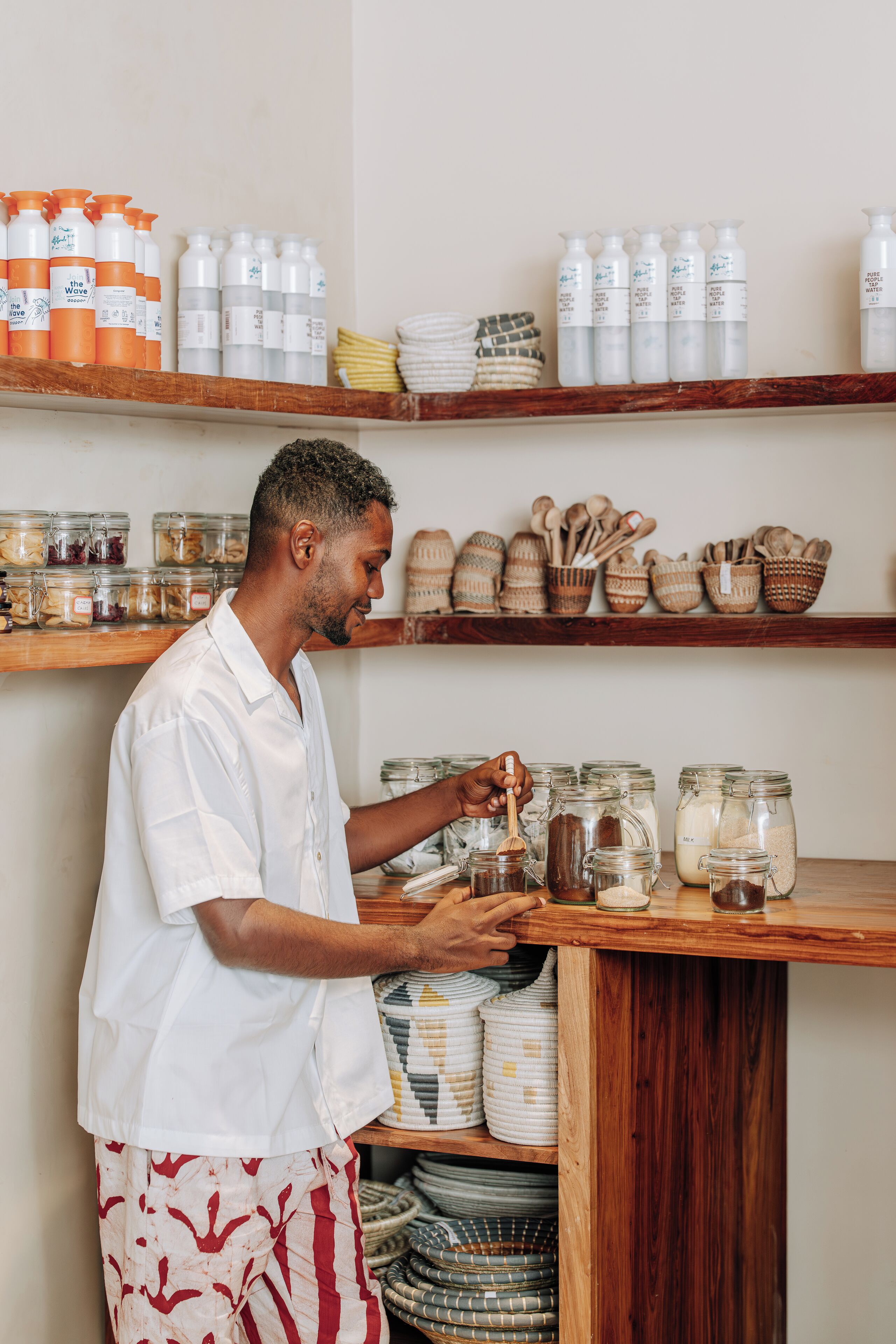 A person in a white shirt and patterned shorts interacting with jars and containers on shelves.