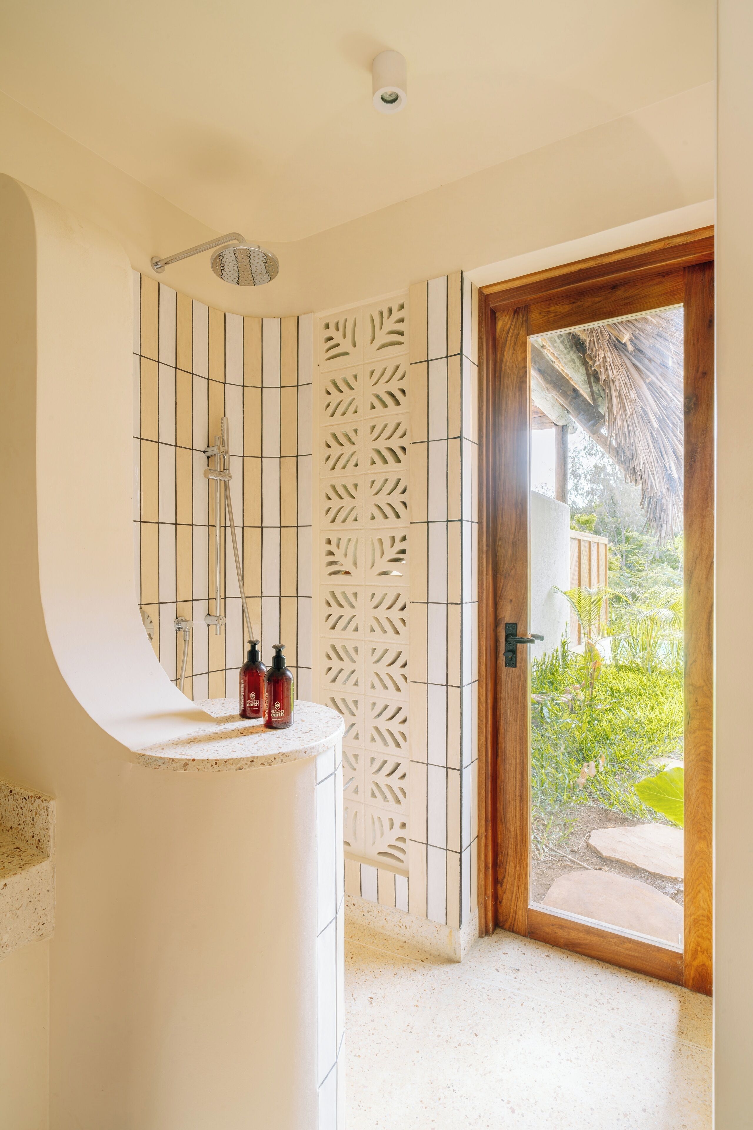 A bathroom with a shower, a toilet, and a sink. There is a decorative wall with a leaf pattern and a wooden door leading to a garden.