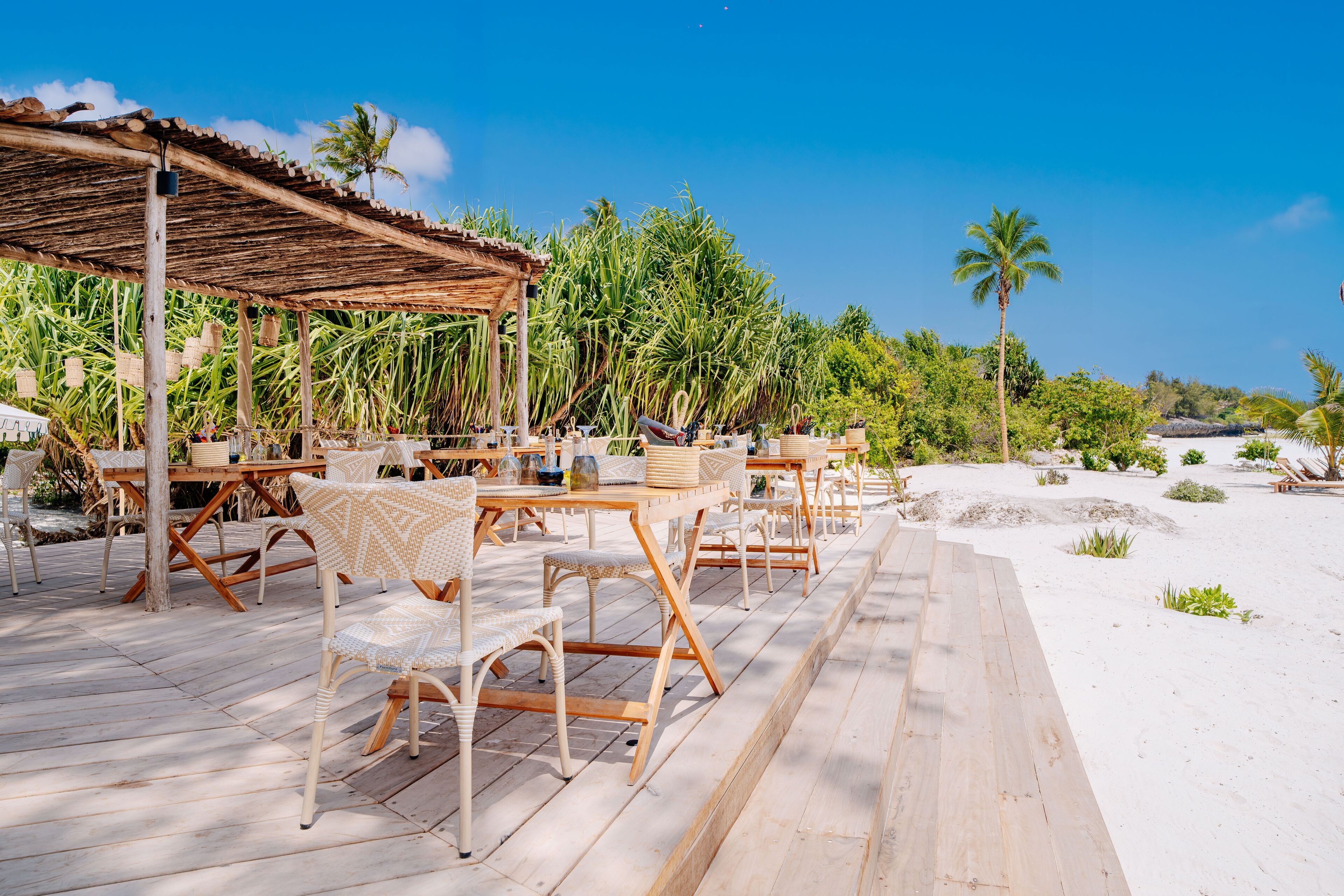 An outdoor dining area with wooden tables and chairs, a covered seating area, and a view of the beach and palm trees.