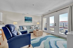 A living room with a sofa, armchair, and coffee table, a painting on the wall, and a view through the glass door. - Adorable 2 bedroom oceanfront condo (North Topsail Beach)