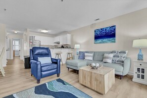 A living room with a blue leather chair, a light blue sofa, a wooden coffee table, and a kitchen in the background. - Adorable 2 bedroom oceanfront condo (North Topsail Beach)