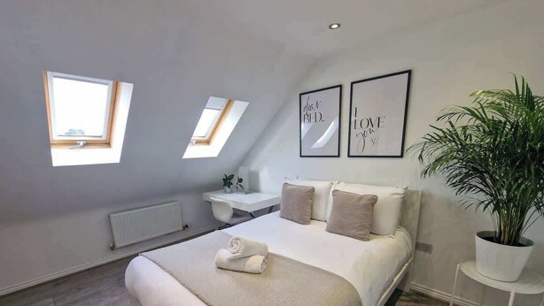 Un dormitorio con una cama, dos mesitas de noche, un radiador, una planta en maceta y dos cuadros en la pared.