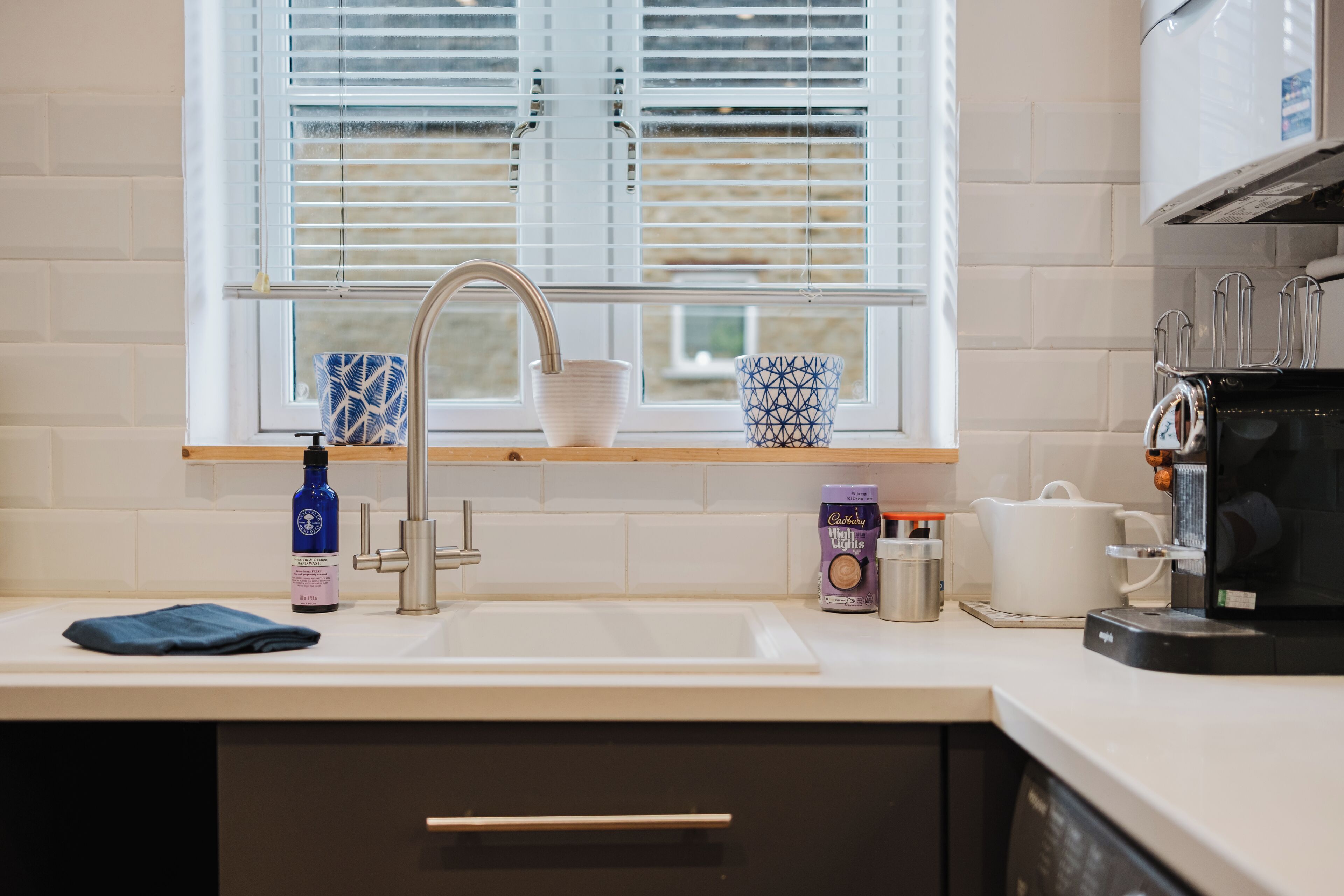 A kitchen with a sink, window blinds, a coffee maker, and a box of Cadbury Dairy Milk.