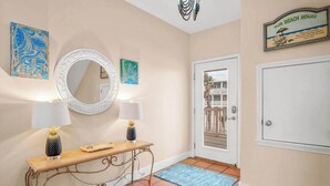 A hallway with a mirror, two wall-mounted lamps, a console table with decorative items, a blue rug, and a door leading to a balcony. - Amazing Beach Views~Boardwalk to Beach~Shared Pool (Port Aransas)