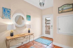Un pasillo con un espejo, dos lámparas de pared, una consola decorada con objetos, un tapete azul y una puerta que da a un balcón.