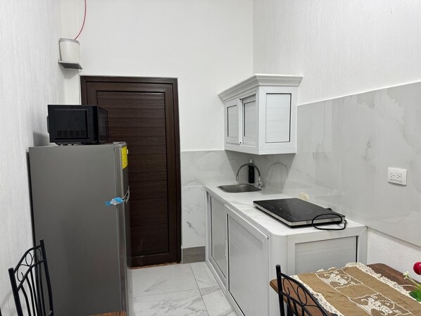 Cocina compacta con refrigerador, microondas y una pequeña encimera con tetera y balanza digital.