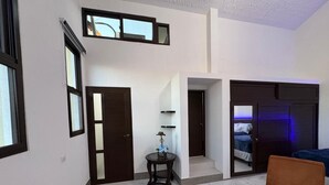 A modern living room with a sofa, a table with a lamp, and a door leading to a bedroom with a bed.