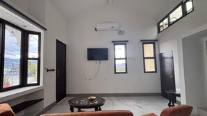 A room with a large window, a flat-screen TV mounted on the wall, a small round table with a decorative item, and two orange chairs.