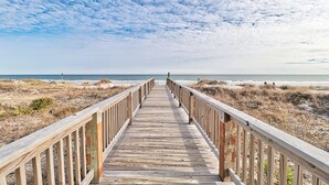 Ein hölzerner Steg führt zu einem Strand unter blauem Himmel.