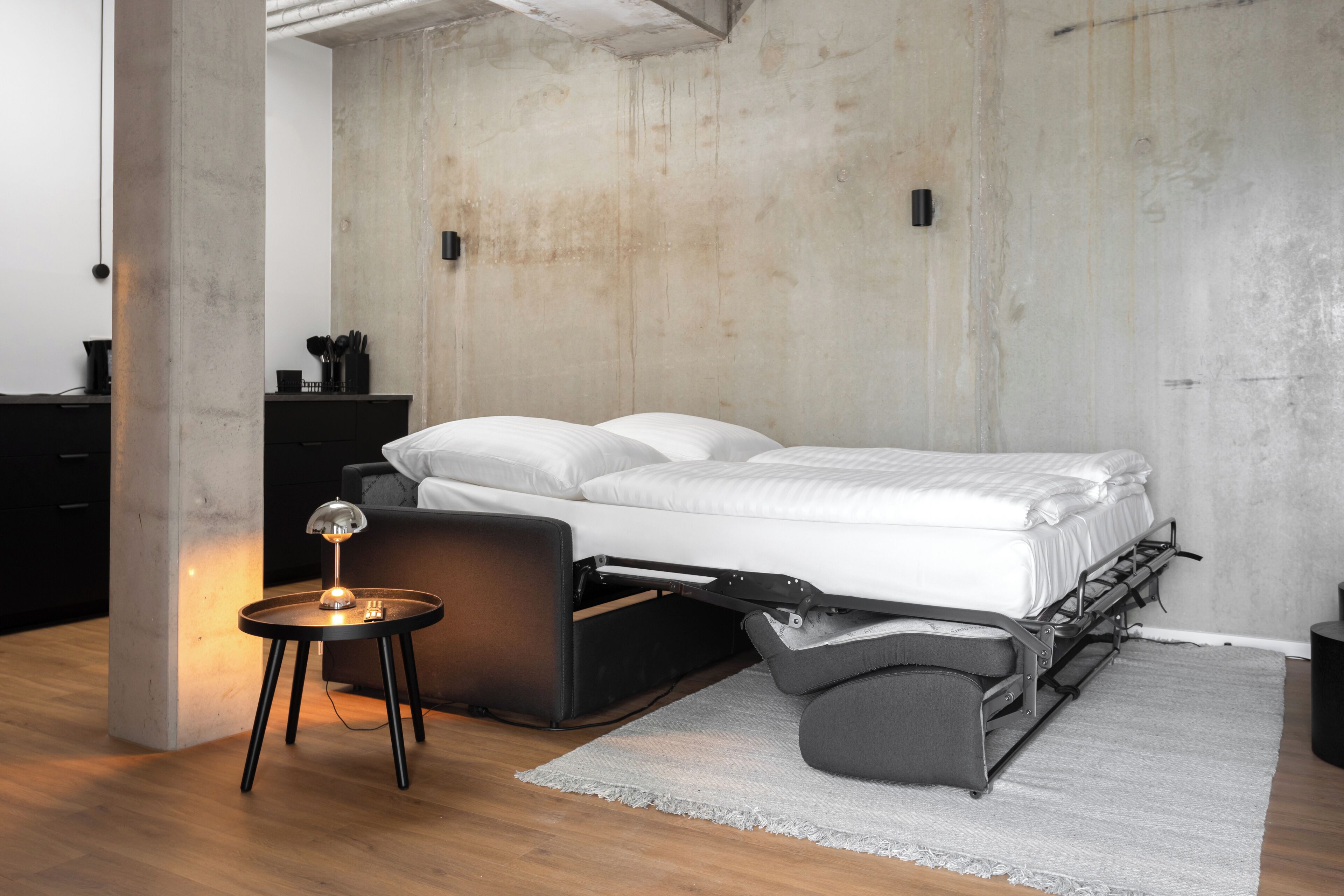Ein modernes Schlafzimmer mit einem Bett, einem Nachttisch mit Lampe und einer Betonwand.