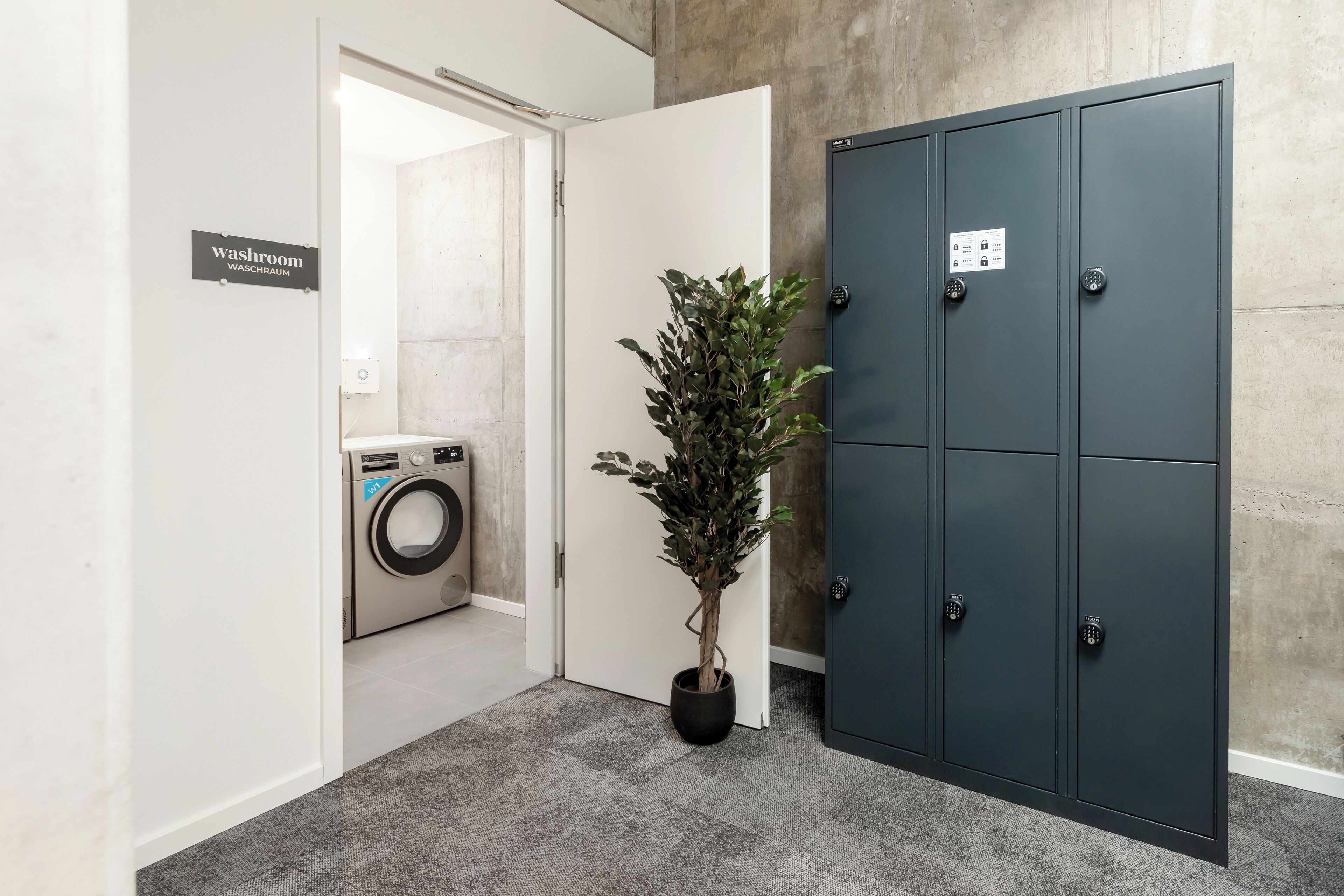 A hallway with a washing machine, a potted plant, and a set of metal lockers.