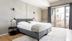 A modern bedroom with a grey bed, a white mattress, and a grey blanket. There is a small black side table with a lamp, and a large window with a view of the outside. - Global Living I The Graf (Berlin)