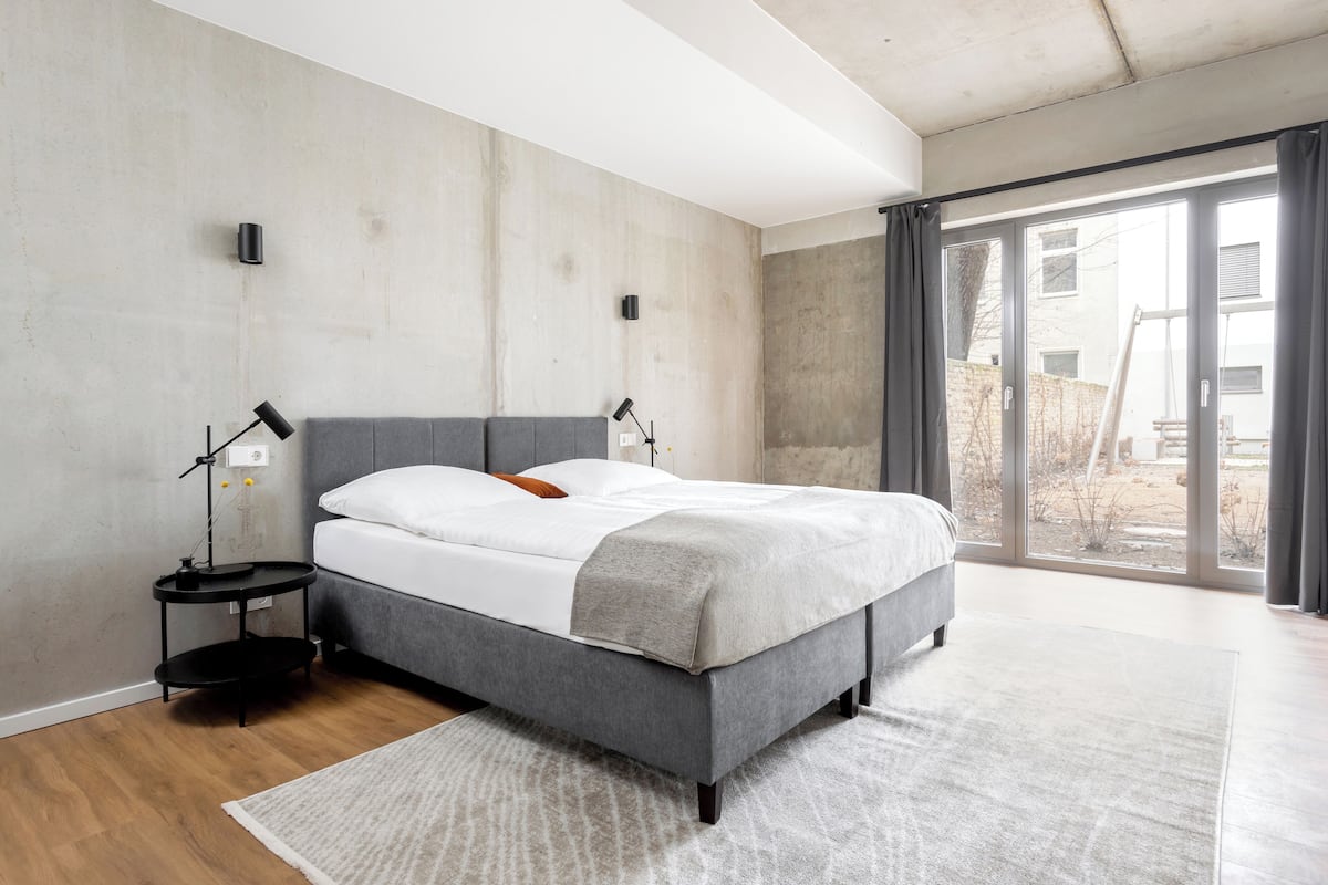 A modern bedroom with a grey bed, a white mattress, and a grey blanket. There is a small black side table with a lamp, and a large window with a view of the outside.