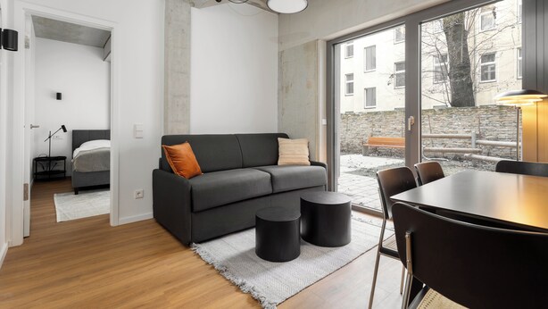 A modern living room with a grey sofa, a dining table, and a large window offering a view of the outside. - Global Living I The Graf (Berlin)